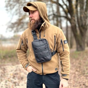 May include: A person wearing a brown fleece jacket with a black crossbody bag. The bag has a zippered pocket and a strap that goes over the shoulder.