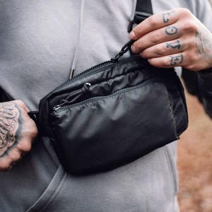 May include: Black crossbody bag with a zippered main compartment and a smaller front pocket. The bag has a black adjustable strap and a sleek, minimalist design. The bag is worn across the chest.