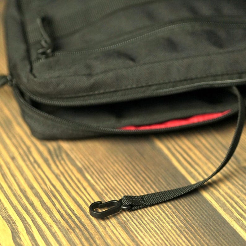 May include: A black zippered bag with a red lining and a black strap with a hook closure.
