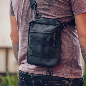 May include: A black crossbody bag with a zippered pocket and adjustable strap. The bag has a tactical look with multiple straps and buckles.