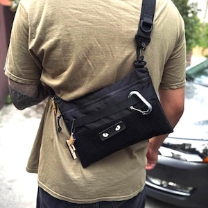 Könnte beinhalten: Schwarze Umhängetasche im taktischen Design mit einem großen silbernen Karabinerhaken und einem schwarzen, verstellbaren Schultergurt. Die Tasche hat eine rechteckige Form mit einem Reißverschluss oben und einem Klettverschluss. Ein kleiner Schlüsselanhänger hängt an der Seite.