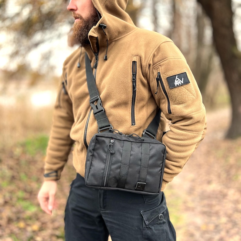 May include: A black crossbody bag with a zippered closure and adjustable strap. The bag is worn by a person wearing a brown fleece jacket with a black zipper and a patch with the text "GRAY" on the sleeve.