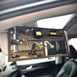 May include: A camouflage car visor organiser with multiple pockets and compartments. It holds various items, including a flashlight, carabiner, pens, and a document. The organiser is attached to the car's visor.