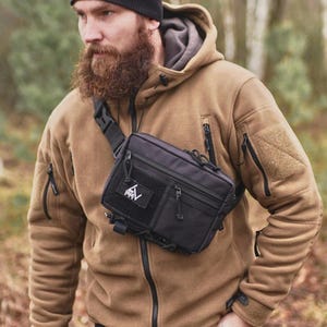 Puede incluir: Una bandolera táctica negra con múltiples compartimentos con cremallera y un parche de velcro. La bolsa se lleva sobre una chaqueta de forro polar color canela. La persona lleva un gorro negro.