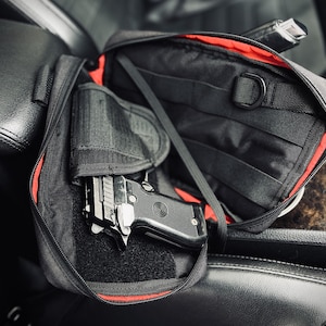 Puede incluir: Funda de pistola de nailon negro con forro interior rojo. La funda está abierta, revelando una pistola negra con corredera plateada. La funda está diseñada para sujetar la pistola de forma segura. La funda está sobre un asiento de cuero negro.