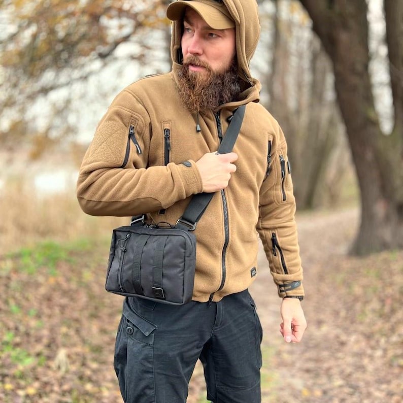 May include: A man wearing a brown fleece jacket with a black crossbody bag. The bag has a zippered pocket and a flap closure.