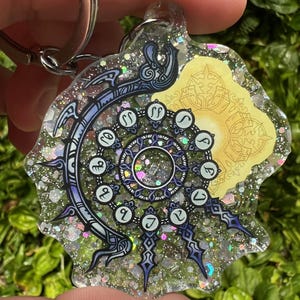 May include: A clear resin keychain with a silver key ring. The keychain features a black and white design of a circle with numbers and a symbol. The background is filled with iridescent glitter.