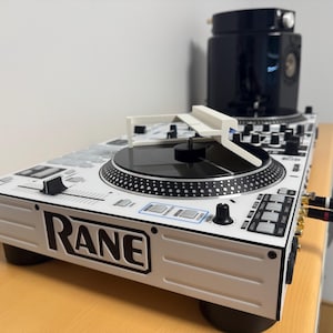 Peut inclure: Une table de mixage DJ Rane blanche et noire avec une platine noire et des boutons de commande. La table de mixage porte le logo Rane à l'avant et est connectée à une enceinte noire. La table de mixage est sur une surface en bois.