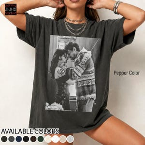 以下が含まれることがあります： 2 人が抱き合っている白黒写真がプリントされたペッパーカラーのTシャツ。「Vintage Vice」と表示されています。シャツはクルーネックと半袖です。複数の色で利用できます。