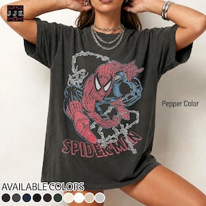 May include: A pepper-colored short-sleeve t-shirt featuring a graphic print of Spider-Man in red and blue with web patterns. The text "SPIDER-MAN" is printed in red below the character. The shirt has a crew neckline and a relaxed fit.