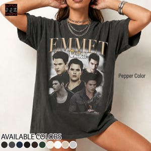 May include: A pepper-coloured short-sleeve t-shirt featuring a vintage-style graphic print of the character Emmet Cullen from the Twilight series. The design includes multiple black and white portraits of the character with the text Emmet Cullen.