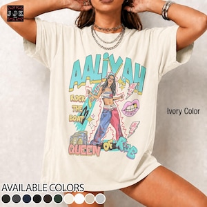 May include: An ivory t-shirt featuring a graphic print of a person in a tube top and split-color pants. The text reads AALIYAH, ROCK THE BOAT, QUEEN OF R&B, and POWER. The design includes a boombox, stars, and a mouth graphic in teal, pink, and yellow.