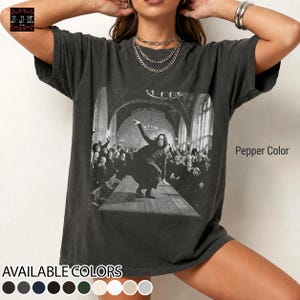 May include: A pepper-colored short-sleeve t-shirt featuring a black and white graphic print of a person in a long coat standing in a large hall with a crowd of people. The shirt has a crew neckline and a loose fit. The text Pepper Color is visible.
