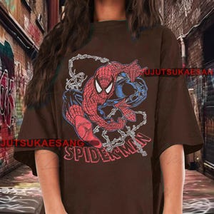 May include: A brown t-shirt featuring a graphic of Spider-Man in red, blue, and black. The word "SPIDER-MAN" is printed in red across the bottom. The design includes web details.