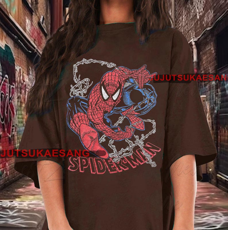 May include: A brown t-shirt featuring a graphic of Spider-Man in red, blue, and black. The word "SPIDER-MAN" is printed in red across the bottom. The design includes web details.