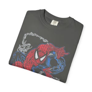 May include: Dark gray t-shirt featuring a graphic of Spider-Man in red and blue, with white web details. The shirt has a crew neck and a small tag on the collar. The design is a vintage comic book style.