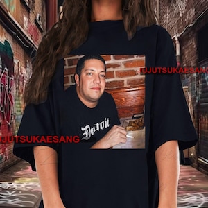 May include: Black t-shirt featuring a framed image of a man with the word "Detroit" in a stylized font. The man is holding a cigarette. The background includes a brick wall and wooden table.