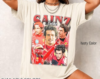 Op de afbeelding: Ivoor kleurig T-shirt met een afbeelding van Carlos Sainz en de rode tekst "SAINZ". Het ontwerp bevat meerdere afbeeldingen van de coureur in een rood racepak. Het shirt toont ook "BESCHIKBARE KLEUREN" met een kleurenpalet.