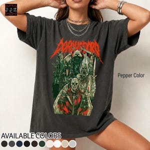 Op de afbeelding: Een peperkleurig T-shirt met korte mouwen en een grafische print van meerdere donkere, skeletachtige figuren en wezens in rood, groen en wit. De tekst "DORHUNDORD" staat in een grillig rood lettertype boven de illustratie.