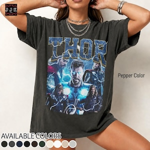 May include: Pepper color t-shirt featuring a graphic of Thor with the word "THOR" in a blue, distressed font. The shirt displays multiple images of Thor. The available colors are shown at the bottom of the image.