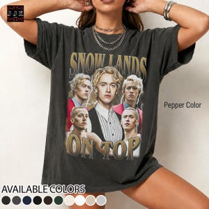 May include: A pepper-coloured short-sleeve t-shirt featuring a graphic print of a person in multiple poses with the text SNOW LANDS ON TOP. The design includes gold-toned lettering and a vintage-style collage of a person in a suit and red jacket.
