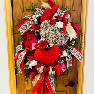 May include: A Valentine's Day wreath with red and gold ribbon, leopard print fabric, and red hearts. The wreath is decorated with white roses and red gems.