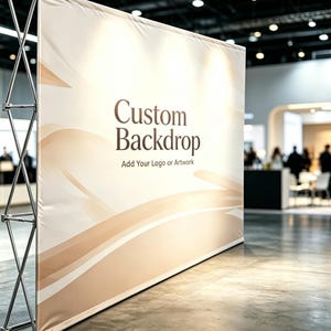 May include: A large, custom backdrop with a cream-coloured background and flowing, light brown design elements. The text "Custom Backdrop" is displayed in a dark brown font, with the tagline "Add Your Logo or Artwork" below. The backdrop is supported by a silver metal frame.