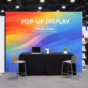 May include: A pop-up display with a vibrant, wavy background. The display reads "POP-UP DISPLAY" and "YOUR LOGO". A black table with a laptop, brochure, and plant is in front of the display. Two grey chairs are in front of the table.