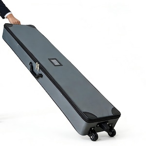 Op de afbeelding: Een grijze, rechthoekige rolkoffer met een zwarte rits, handvat en hoekbeschermers. De koffer heeft een klein, rechthoekig, helder venster en wordt getrokken. De koffer heeft vier wielen.