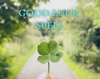 May include: A four-leaf clover, a symbol of good luck, held in a hand against a blurred background of green foliage. The text "GOOD LUCK SPELL" is written in blue above the clover.