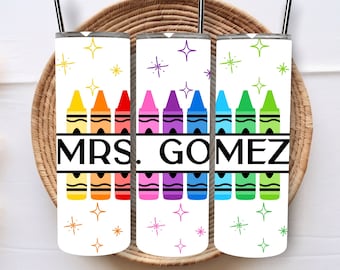 Puede incluir: Tres vasos blancos con un arcoíris de lápices de colores y el texto "MRS. GOMEZ" impreso en ellos. Los vasos están sobre un posavasos de mimbre marrón.