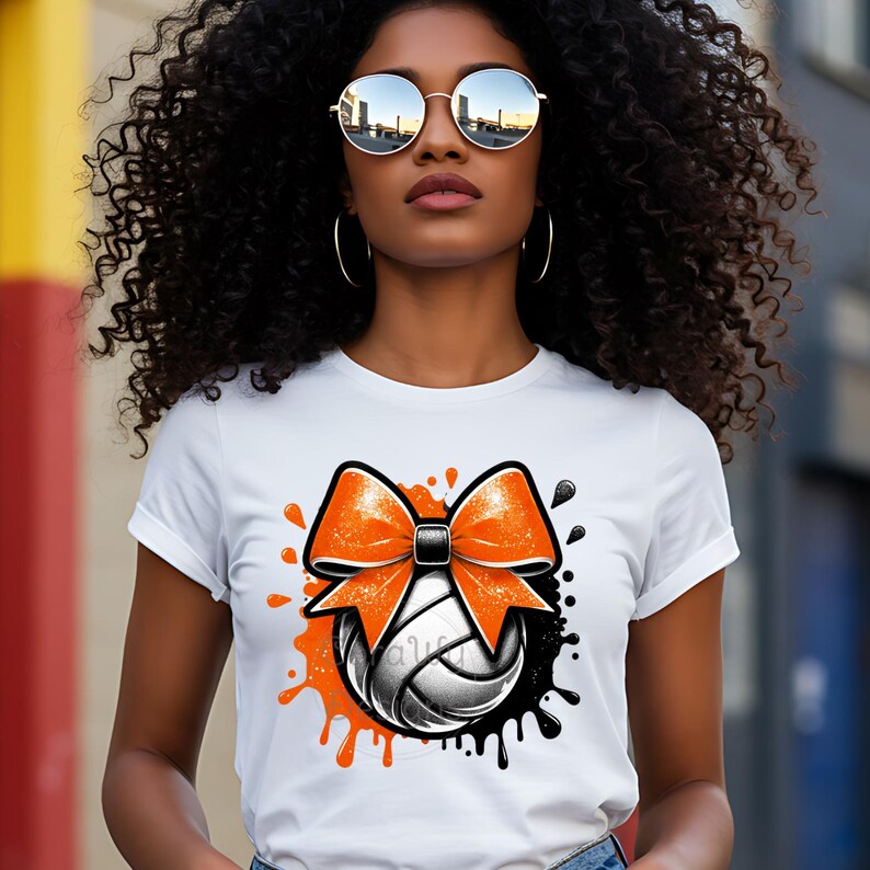 May include: White t-shirt featuring a volleyball design with an orange bow and black and orange paint splatters. The volleyball is white and gray. The shirt is a crew neck.