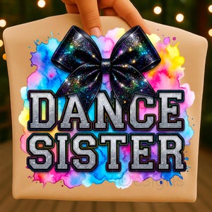 May include: A tan-colored pillow with a large black bow featuring a glittery design. The pillow has the words "DANCE SISTER" in silver block letters with a black outline. A colorful watercolor splash design is behind the text and bow.