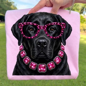 May include: A light pink t-shirt featuring a graphic illustration of a black Labrador retriever wearing bright pink leopard print glasses and a necklace made of large, square, faceted hot pink gemstones set in silver-toned metal frames.