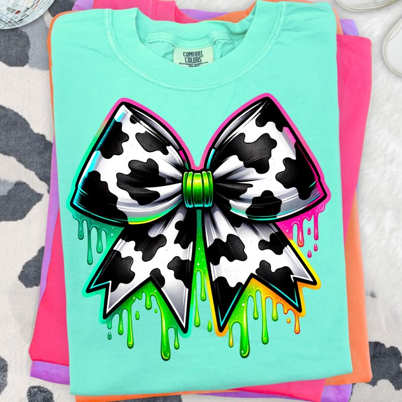 May include: A mint green t-shirt featuring a large graphic of a bow with a black and white cow print pattern. The bow has a bright green center knot and is surrounded by vibrant neon green and orange paint drip effects at the bottom edges.