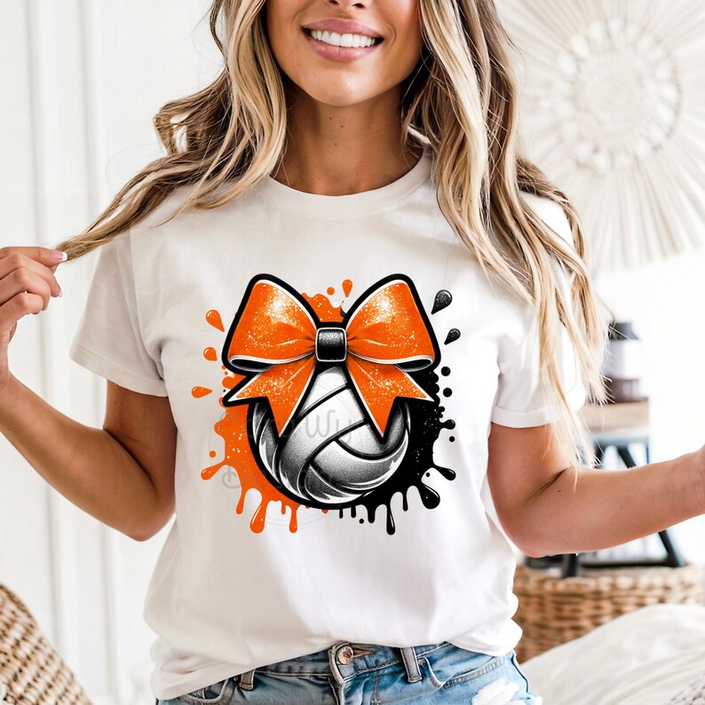 May include: White t-shirt featuring a volleyball design with an orange bow and black accents. The volleyball is white and gray, with orange and black paint splatters around it. The design is centered on the front of the shirt.