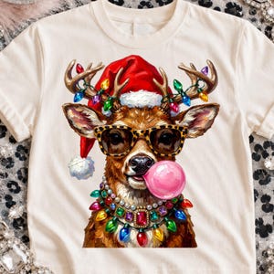 May include: A cream-colored t-shirt featuring a graphic of a reindeer wearing a red Santa hat, leopard print glasses, and a necklace made of colorful Christmas lights and pearls. The reindeer is blowing a large, round, bright pink bubble.