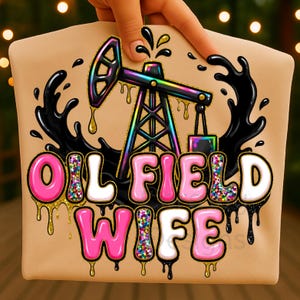 May include: Tan t-shirt featuring a colorful oil rig graphic with the words "OIL FIELD WIFE" in pink and white letters. Black oil-like drips and accents surround the design. The shirt is held by a hand.