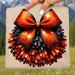May include: A beige t-shirt featuring a graphic of a large, round pom-pom made of orange and black fringe strips. A prominent, glittery orange bow with black edges sits at the top of the pom-pom, adding a decorative element to the design.
