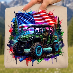 Può includere: Una felpa beige con una grafica vibrante. Raffigura un ATV verde con ruote blu luminose, guidato da una persona con un casco, sullo sfondo di una bandiera americana, alberi e schizzi di vernice colorati.
