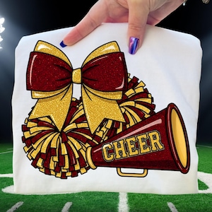 May include: White t-shirt featuring a cheerleading design. The design includes a large maroon and gold glitter bow, maroon and gold pom-poms, and a maroon megaphone with the word "CHEER" in gold lettering. The shirt is held by a hand.