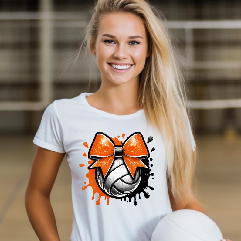 May include: White t-shirt featuring a volleyball design with an orange bow and black accents. The volleyball is black and white. The design includes orange paint splatters. The shirt is a casual style.