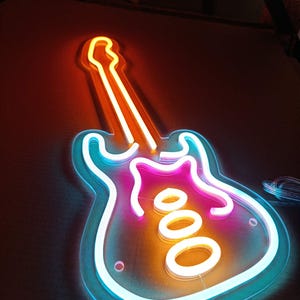 Peut inclure: Une enseigne de guitare au néon aux couleurs vives. Le contour est bleu vif, avec un centre rose et orange. Le manche de la guitare est orange et blanc. L'enseigne est conçue comme un élément décoratif.