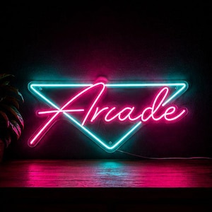 Peut inclure: Enseigne au néon avec le mot "Arcade" en rose, superposé à un triangle turquoise. L'enseigne est fixée sur une surface sombre, avec un fond sombre. L'enseigne est allumée, créant une esthétique rétro.