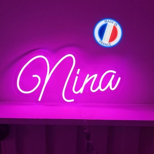 Peut inclure: Enseigne au néon rose vif avec le nom "Nina" écrit en cursive. Un autocollant "Made in France" est visible. L'enseigne est posée sur une étagère blanche sur fond violet. Un objet décoratif.