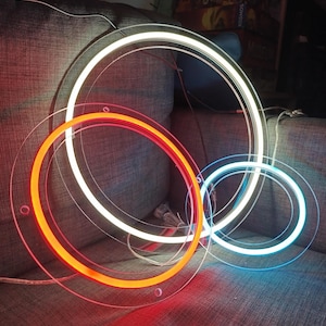 Peut inclure: Une œuvre d'art moderne avec trois luminaires circulaires au néon. Les lumières sont rouges, blanches et bleues, avec des supports en acrylique transparent. L'arrangement est sur une surface grise.
