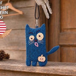 May include: A dark blue crocheted cat keychain featuring large, mismatched eyes, a pink tongue, and a small wooden pumpkin-shaped button attached to the lower body. The text PDF CROCHET PATTERN ENG and a United States flag icon are visible.