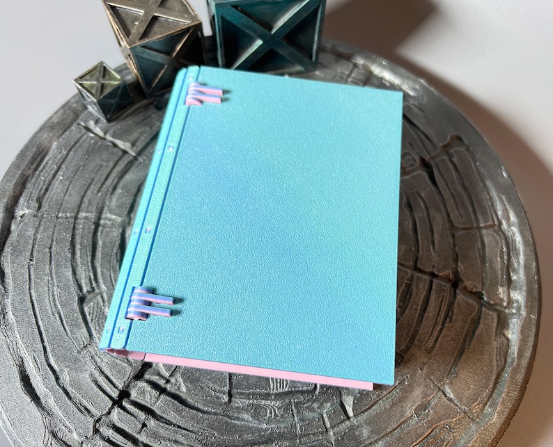 May include: A light blue notebook with pink edges and a textured cover. The notebook is closed and has a series of metal rings along the spine.