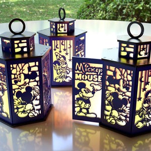 May include: Four dark blue, hexagonal lanterns with black tops and handles. The lanterns feature cut-out designs of Mickey Mouse and friends, with the words "Mickey Mouse" visible on one. The lanterns are illuminated from within.