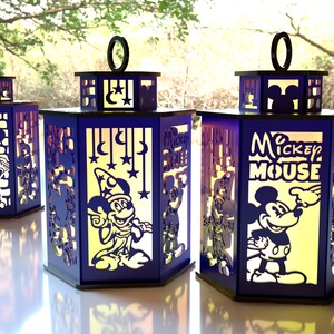 May include: Three blue decorative lanterns with cut-out designs featuring Mickey Mouse and other Disney characters. Each lantern has a black handle and a yellow interior, creating a warm glow. The lanterns are placed on a white surface.